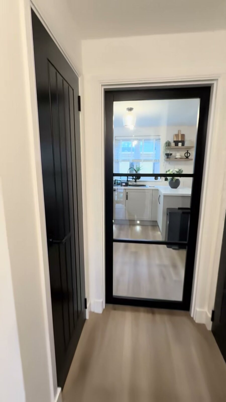 A black Camden glazed door at the end of a hall, into a bright kitchen