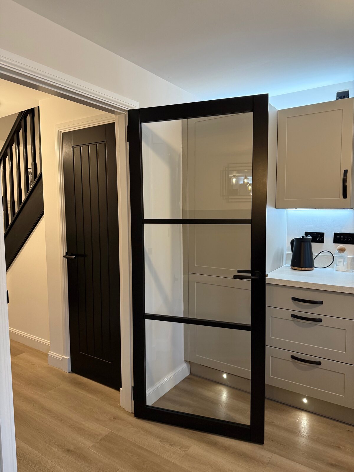 A black urban glazed door open into a kitchen, with a black cottage door and a black-spindle staircase alongside